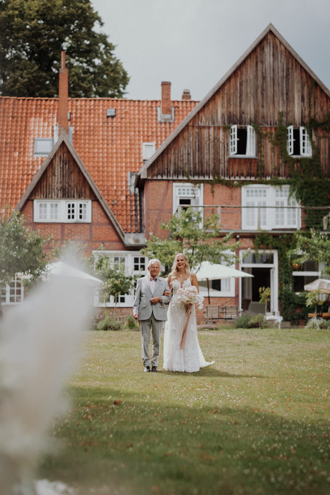 Cyana und ihr Vater unterwegs zum freien Altar, das Hotel im Hintergrund, Pampasgras unscharf im Vordergrund.