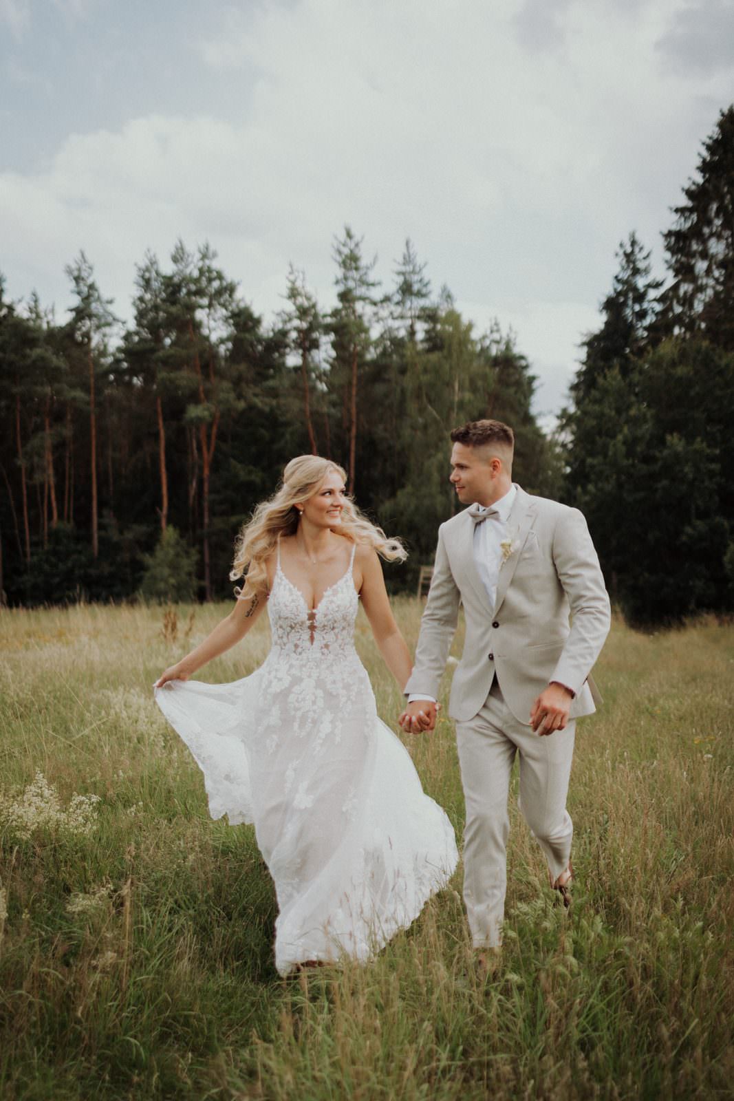 Cyana und Basti Hand in Hand über die Waldwiese — Schleppe im Wind, Kiefernwald im Hintergrund.