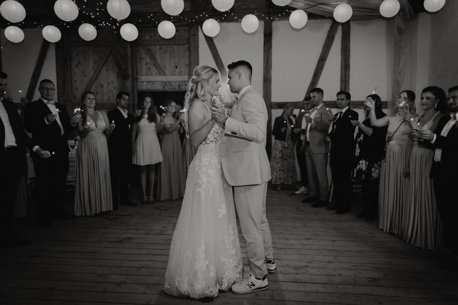 First Dance unter Lampions und Lichterkette — Schwarzweiß, Gäste halten Wunderkerzen am Rand der Tanzfläche.